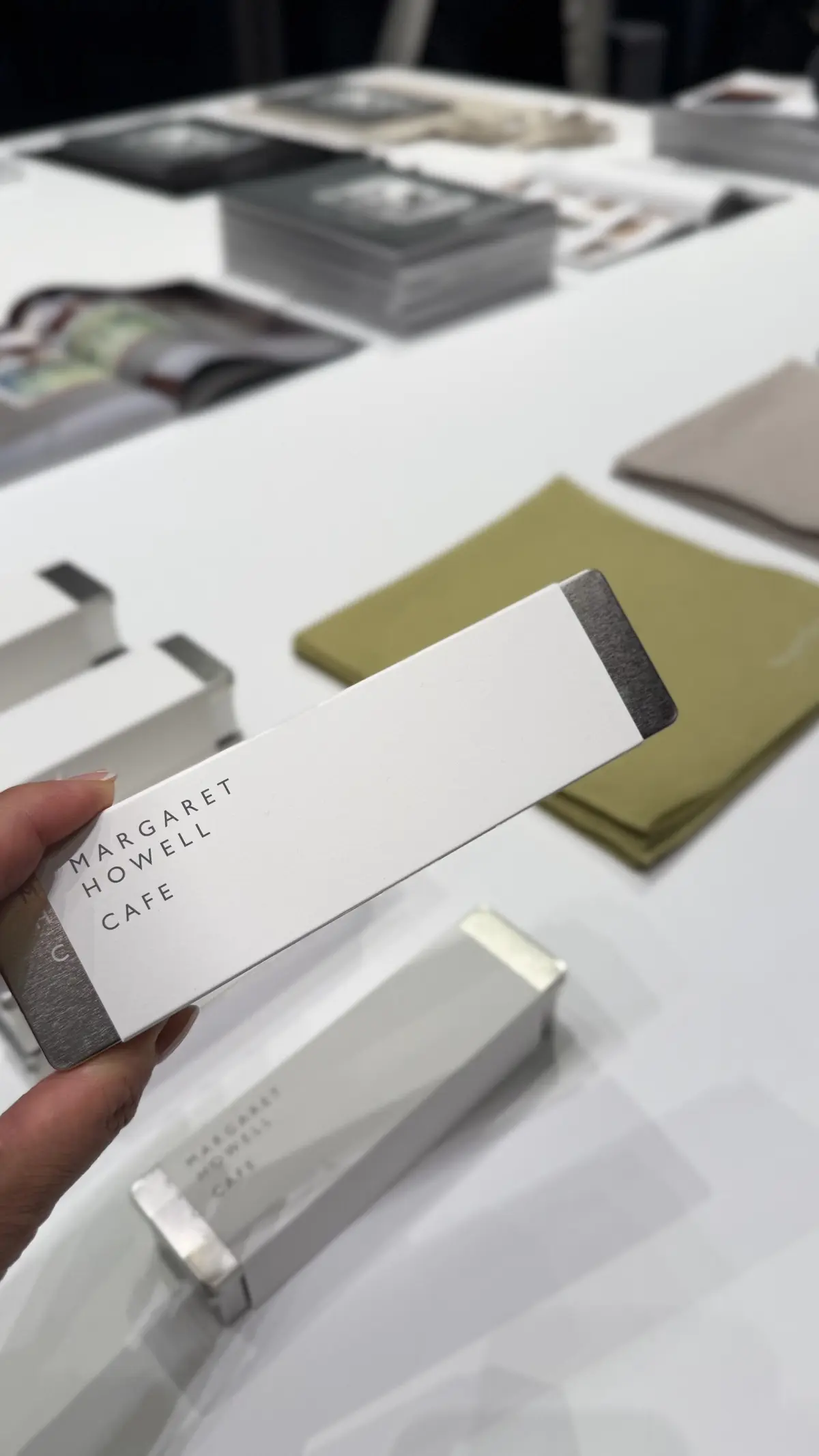 白いテーブルに置かれた、Margaret Howell Cafeのギフトボックス。長方形で、シルバーのロゴがプリントされている。手で保持しているボックスと、テーブルの周りに積み重ねられた同様のボックスが写っている。