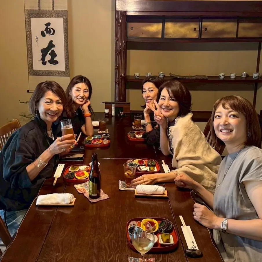 5人の女性が木製テーブルに座り、料理と飲み物を楽しんでいる。各人が異なる料理の盛り合わせとビール、ワインを手に持っている。背景には和風の壁画と棚があり、温かみのある雰囲気。