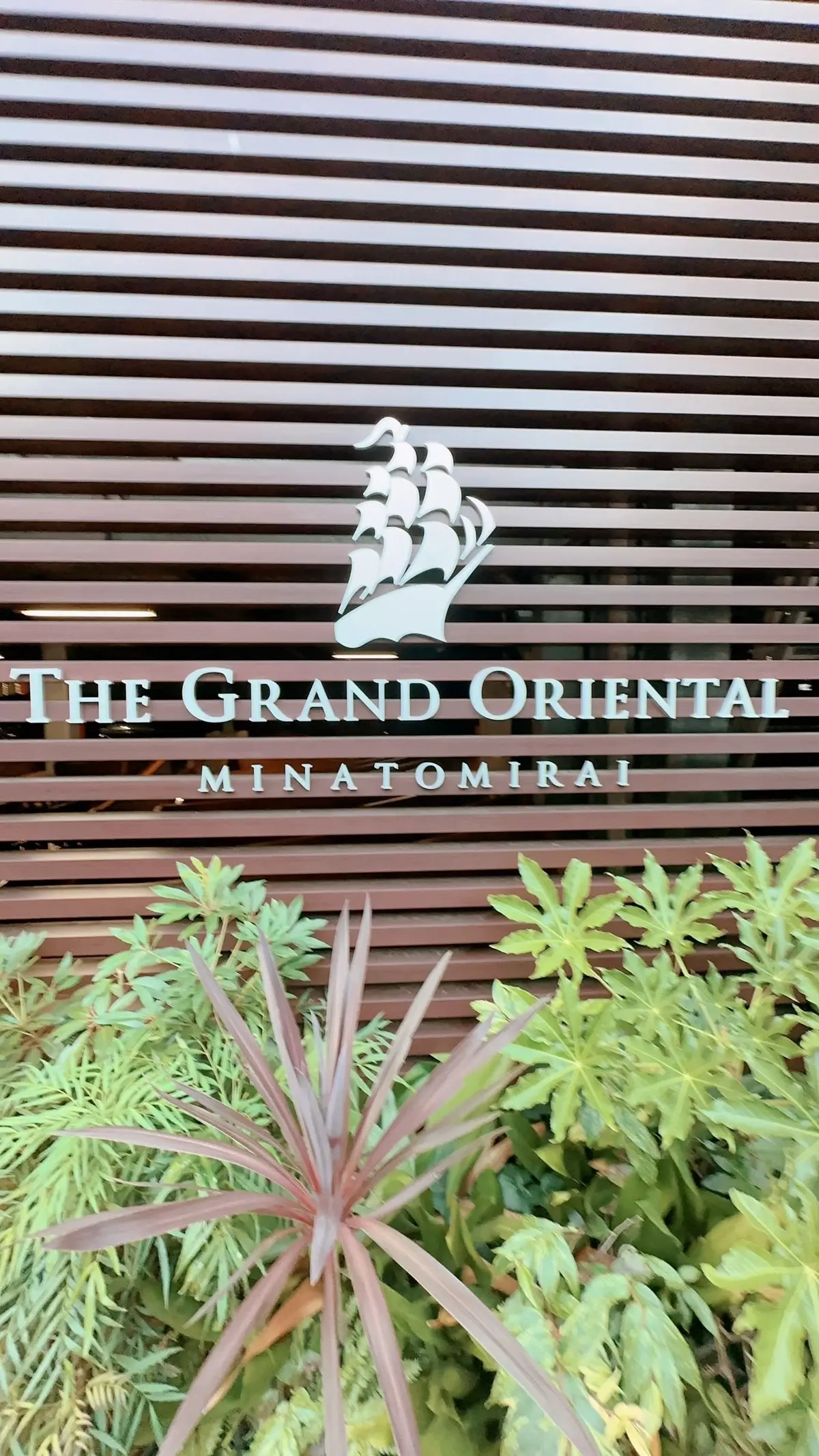 木製の水平スラットフェンスに、The Grand Oriental Minatomirai のロゴマークと文字が刻まれている。フェンスの下には緑の植物が生い茂っている。