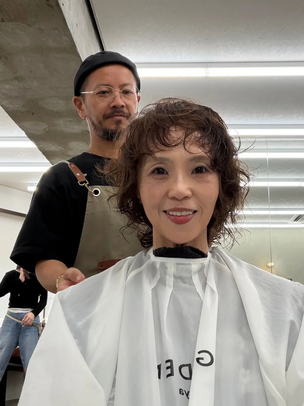 美容師にカットされた女性。茶色のくせ毛で、白いエプロンを着用。背景には、男性の美容師と、コンクリート打ちっぱなしの壁が見える。