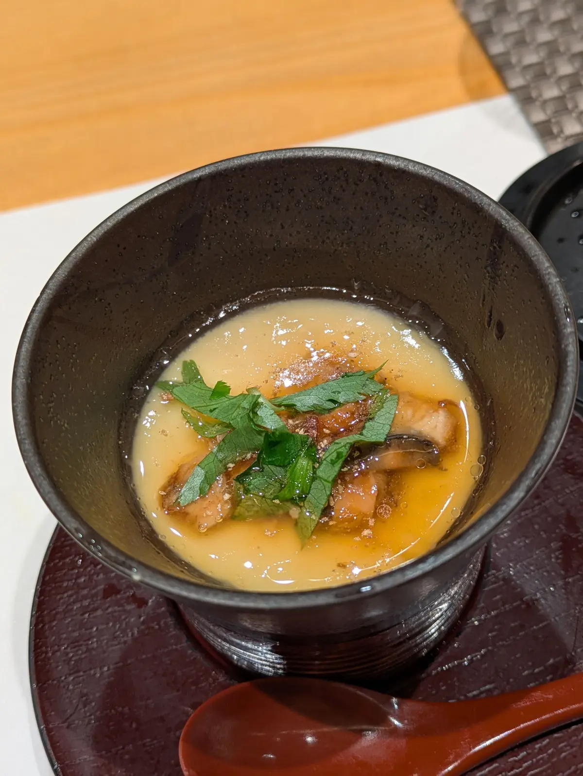 黒い鉢に黄色のスープ、きのことパセリの葉が乗った料理、赤いスプーンが横に置かれている
