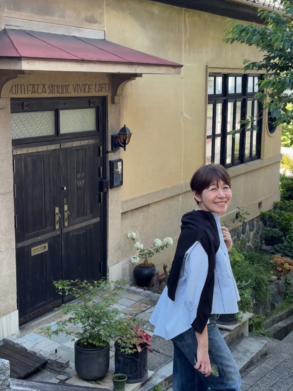ベージュの建物の入り口前で、笑顔の女性が立っている。黒いドアと窓、赤い屋根の軒先、植えられた花や低木が周囲を飾る。