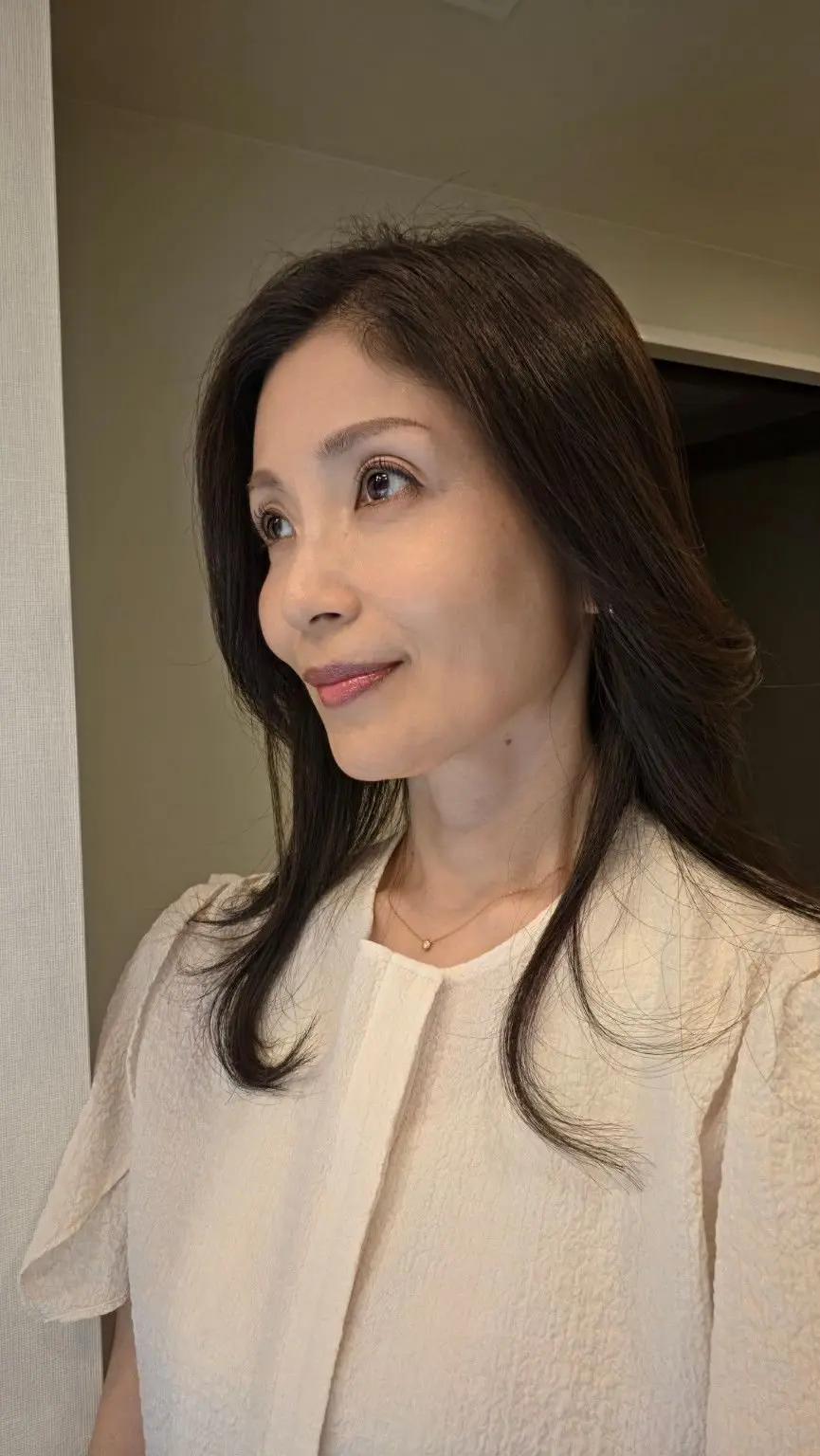 白い襟付きブラウスを着た女性が、やや上を向いて微笑んでいる。茶色の長い髪を肩まで伸ばし、シンプルなネックレスを着用。背景はベージュ色の壁。