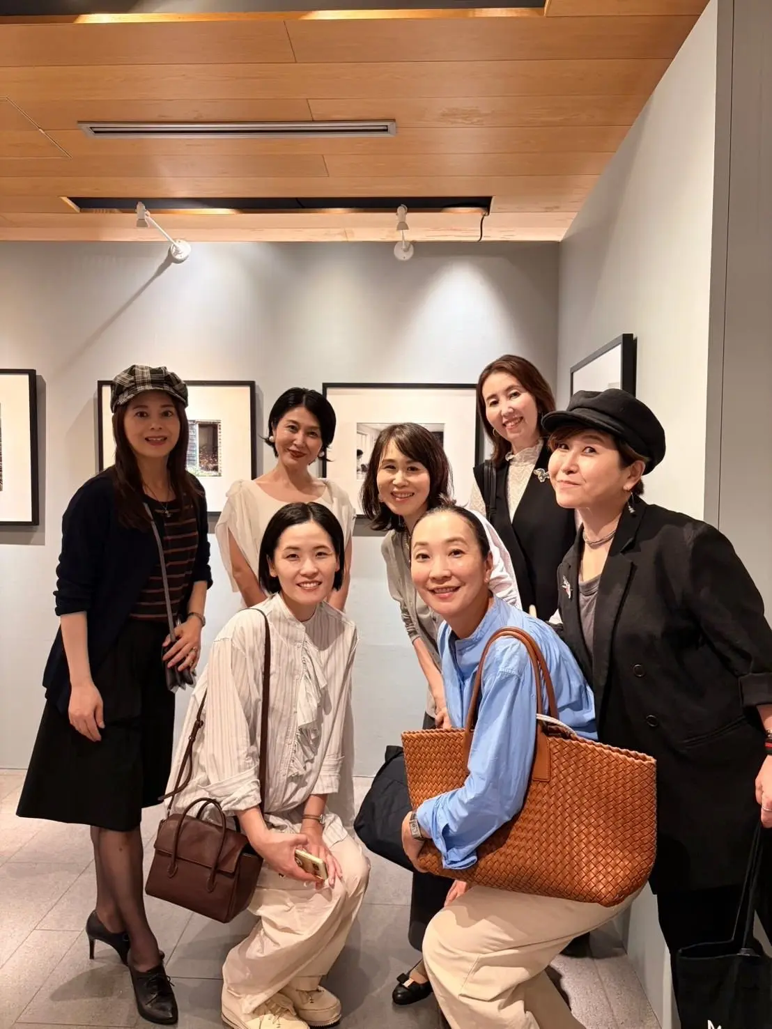 美術館のような空間で、7人の女性が笑顔で写っている集合写真。左から黒い服の女性、チェック柄帽子を被った女性、白いトップスにベージュのパンツ姿の女性、茶色の服の女性、グレーの服の女性、黒いジャケットに帽子を被った女性。背景には白黒の絵画が飾られている。