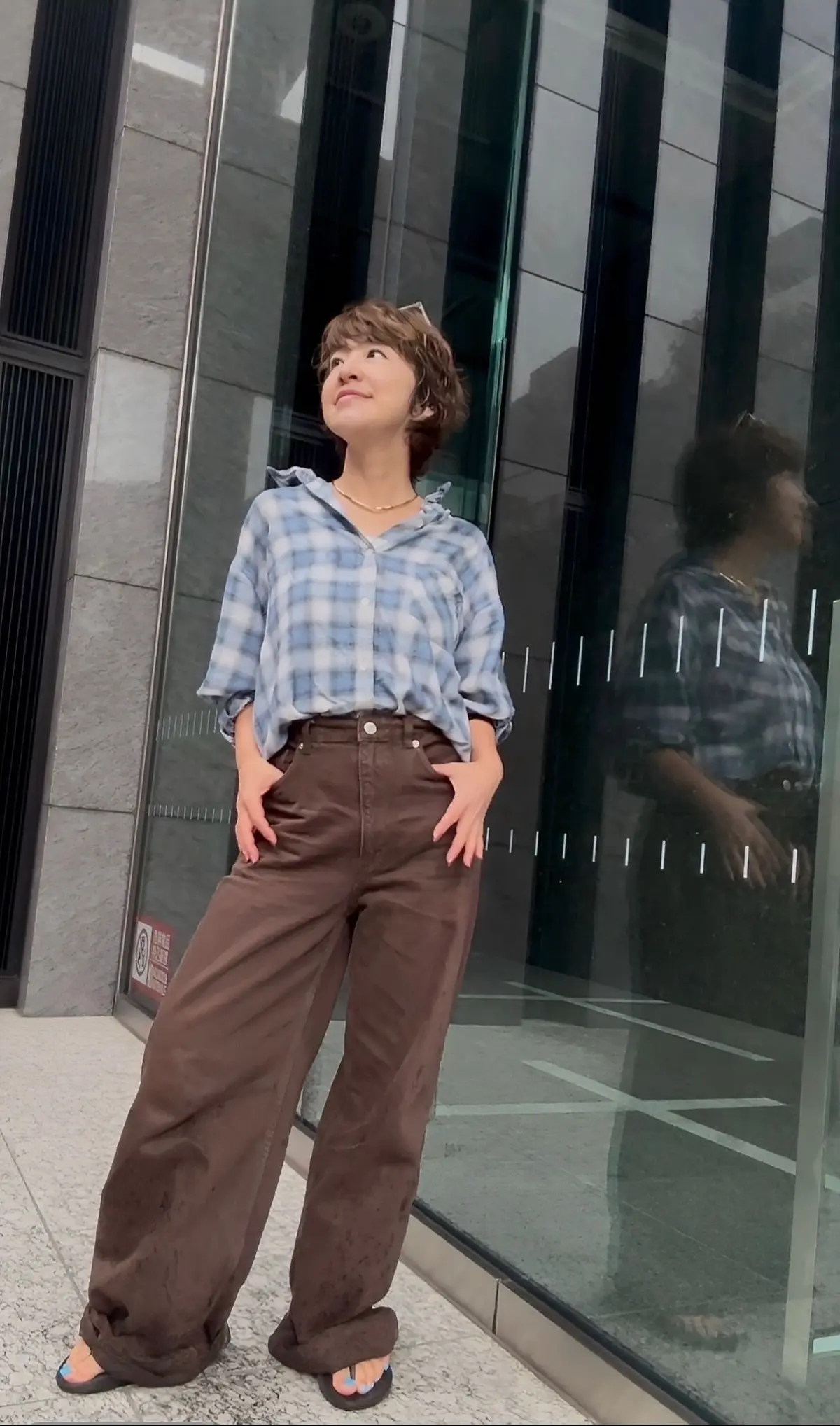 茶色のワイドパンツに青いチェック柄シャツを合わせた女性。短くカットした茶色の髪で、窓際に立って上を見上げている。足元はサンダル。