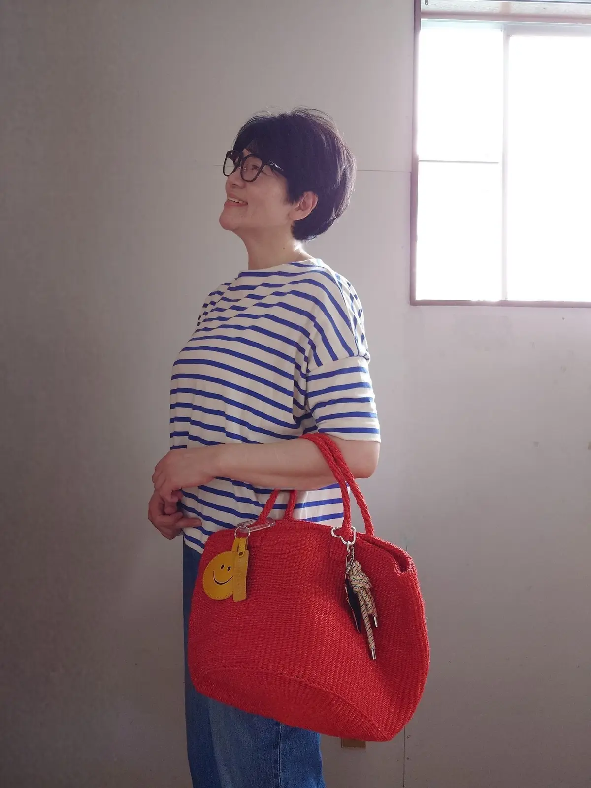 白い線が青色で描かれたボーダーTシャツを着た女性が、赤い編み込みバッグを持って立っている。女性は眼鏡をかけ、微笑んでいる。背景には窓がある。
