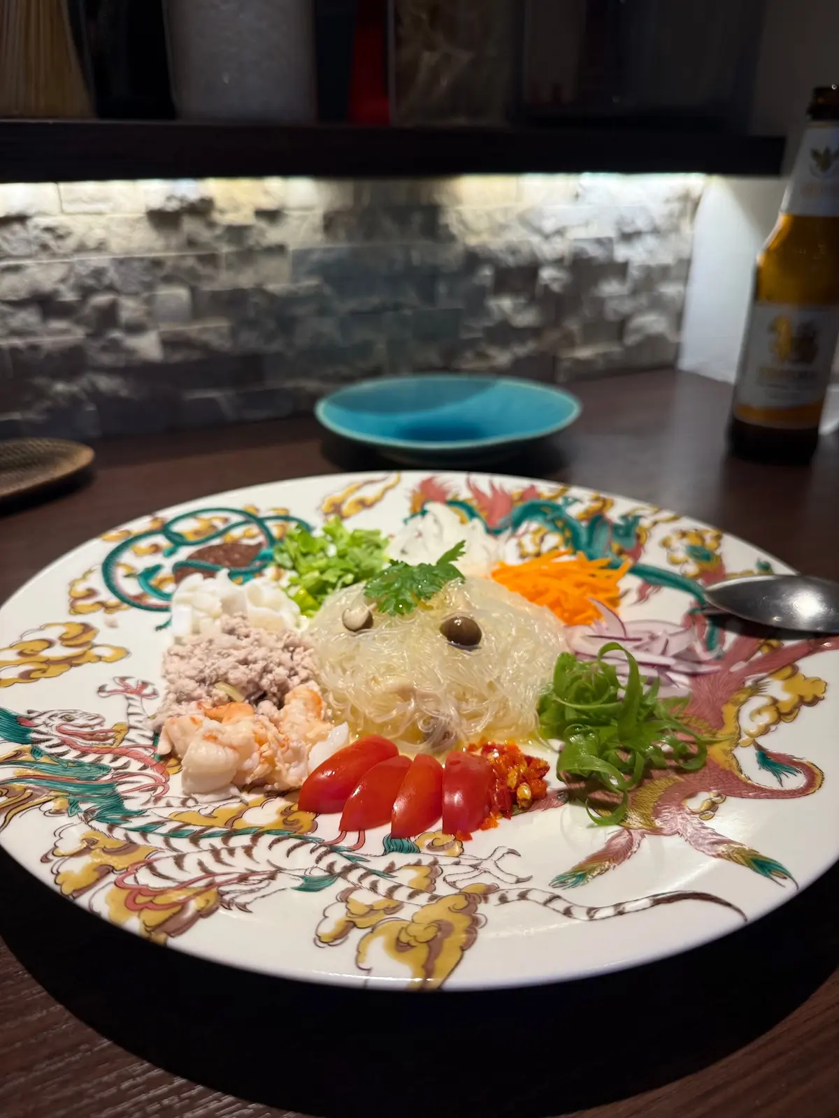 大阪・福島の「Modern Thai CIEL 大阪本店」でいただいた本格タイ料理。縁起の良い龍の絵柄が描かれた皿に、ユースフル（冬虫夏草）の盛り合わせ。白玉、エビ、トマト、ガラス麺、野菜などが彩り豊かに配置されている。背景にはビール瓶と石積みの壁が見える。