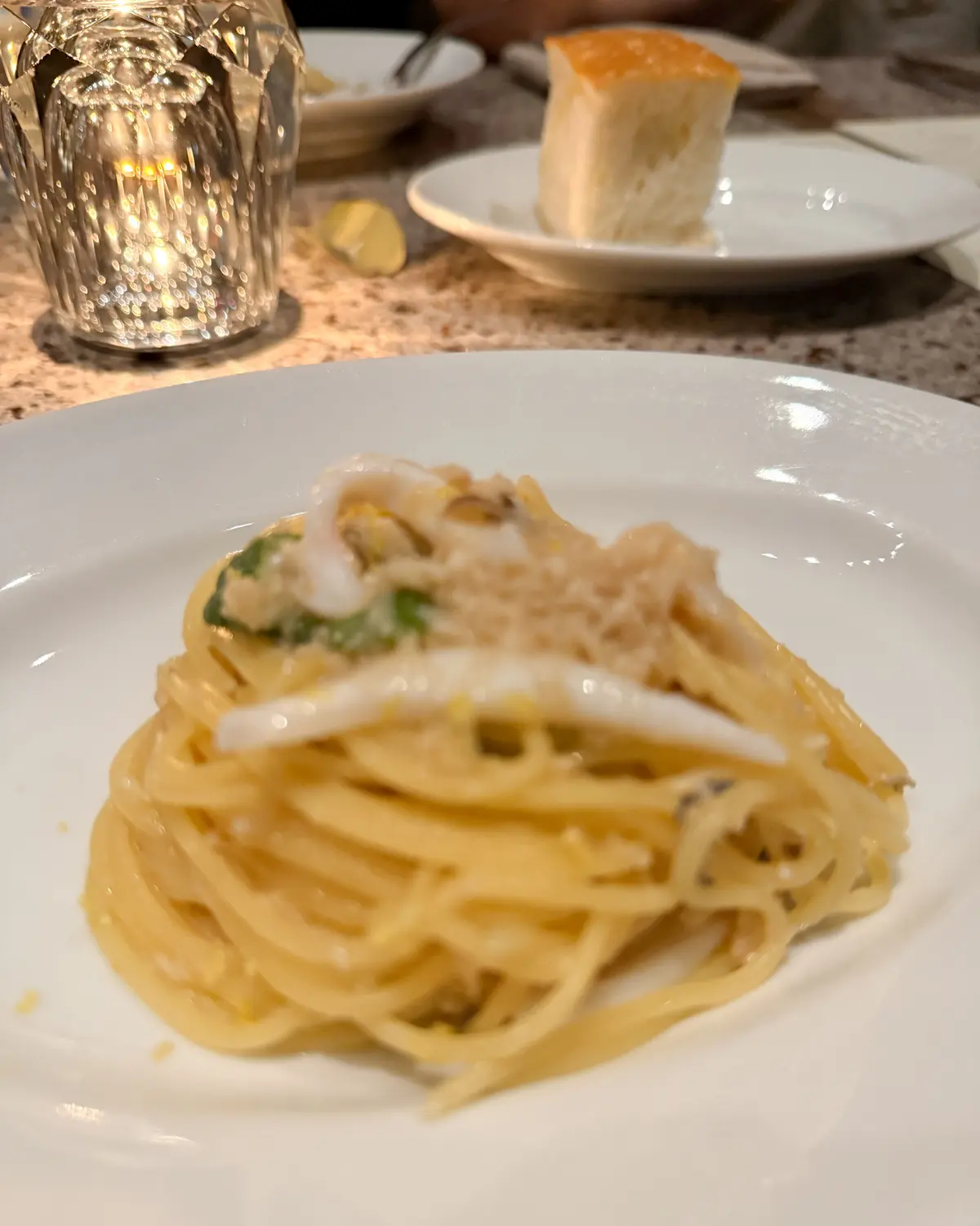 白磁の皿に盛り付けられた、スパゲッティと魚介のパスタ。表面にはチーズが散りばめられ、緑色の葉も見える。背景にはグラスとパンが置かれている。