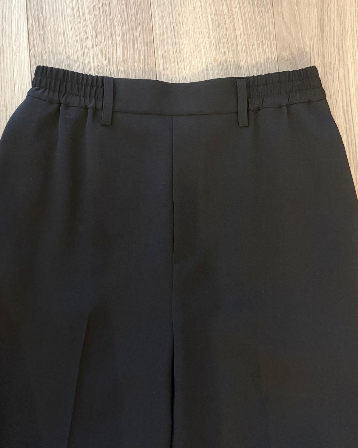 黒いテーパードパンツ。ウエストはゴムで、ボタンが2つ付いている。木製の背景に置かれている。