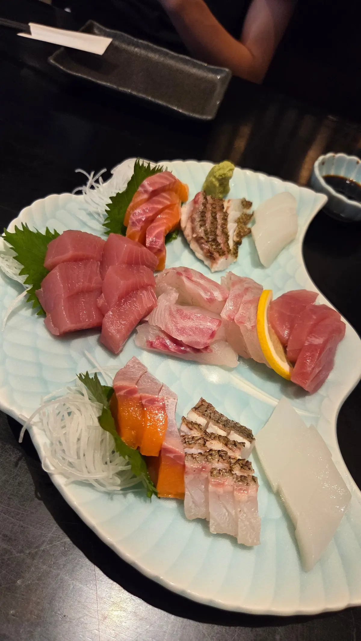 青い皿に盛り付けられた刺身の盛り合わせ。上段にはマグロ、サーモン、ホタテ、カマス。下段にはマグロ、ブリ、ホタテ、カマスが並び、それぞれに柑橘類や薬味が添えられている。右には醤油小皿。