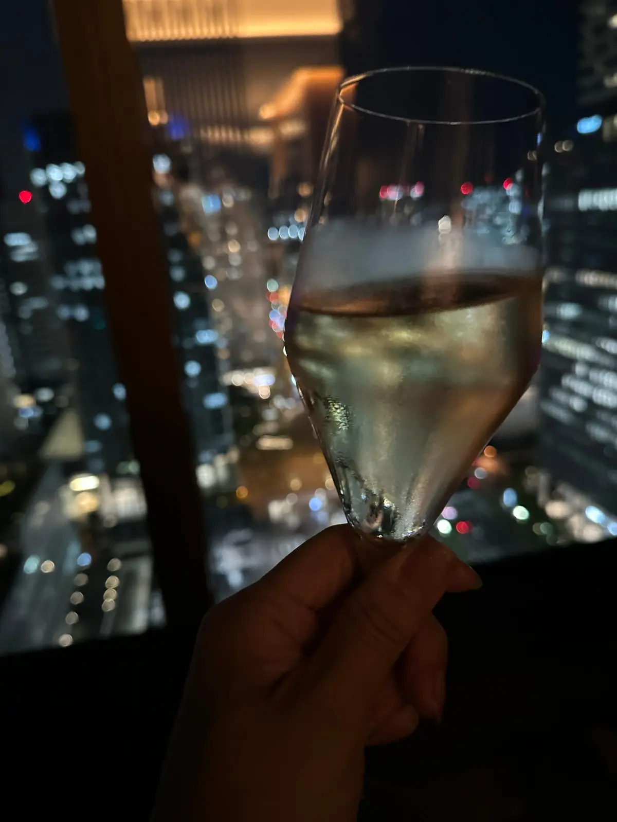 手はワイングラスを保持し、グラスには白ワインが注がれている。背景には夜の街の高層ビルが点在し、街灯やネオンの光がぼやけて輝いている。