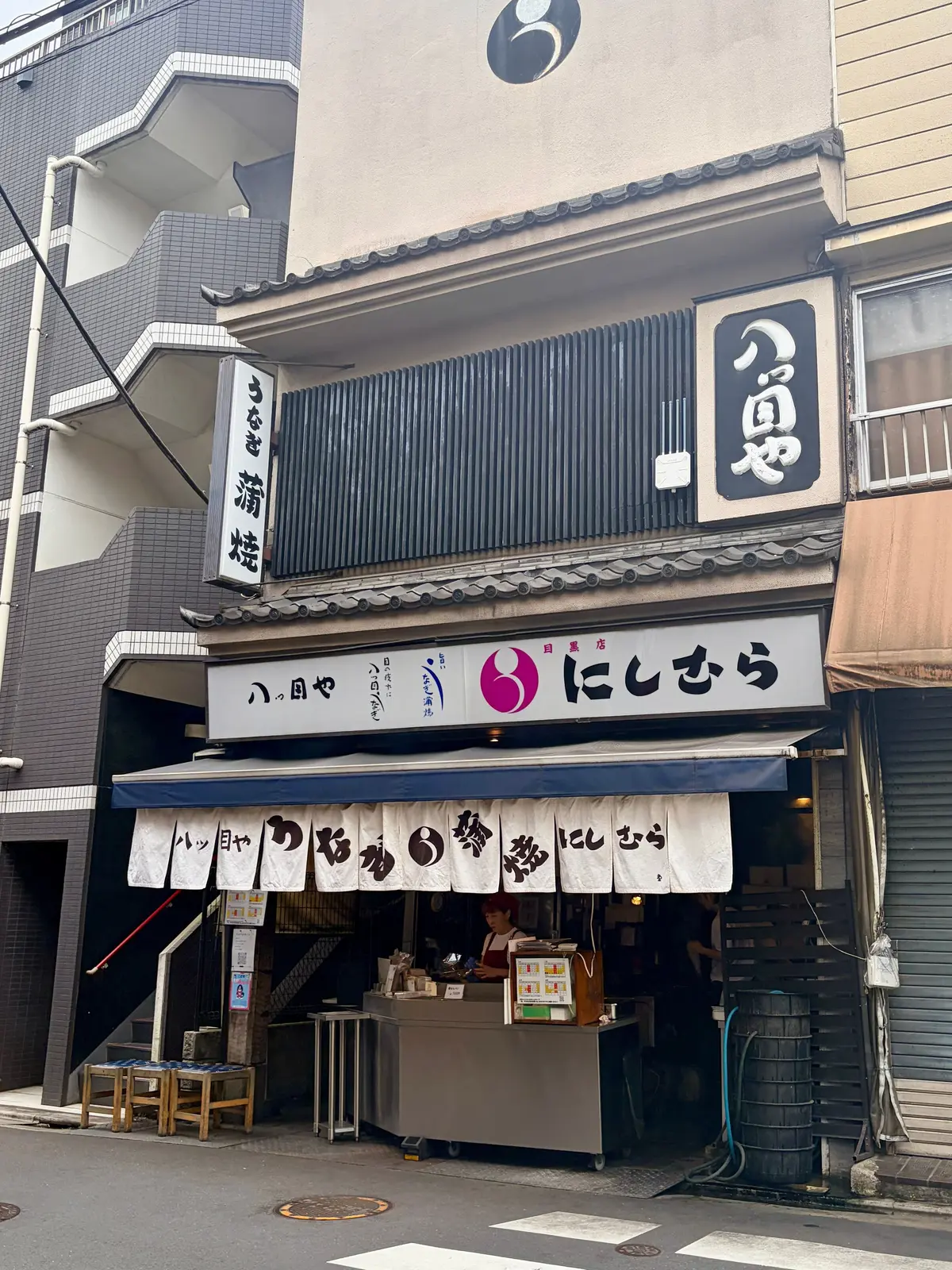 日本の路地裏に佇む、看板が並ぶ「にしむら」という名の飲食店。木造の建物で、格子窓や垂れ幕が伝統的な雰囲気を醸し出している。入り口にはメニューが掲示され、店員が客を待っている。