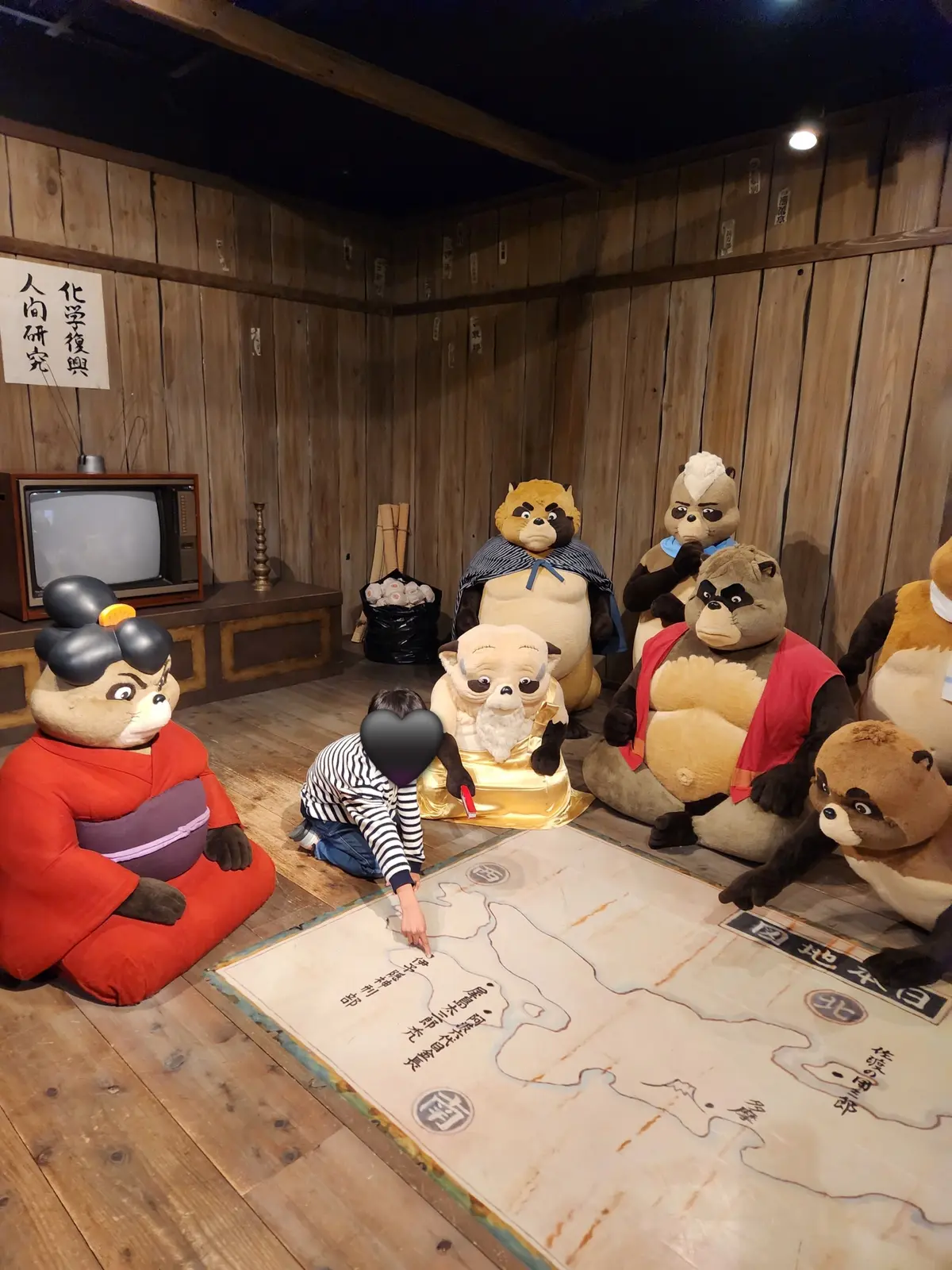 木製の壁の前に、猿の着ぐるみを着た人物が、日本地図が描かれたテーブルを囲んでいる。中央には金色の像が置かれ、一人が触れている。背景には古いテレビと、漢字表記の看板がある。