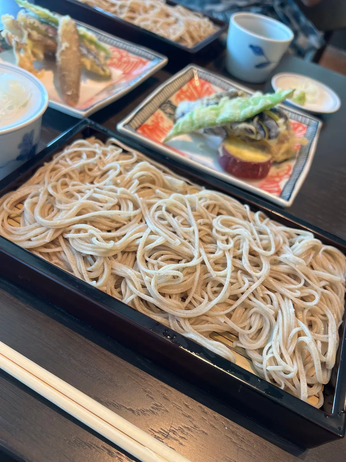 白いうどんが黒い長方形の皿に盛り付けられ、周囲には刺身、焼き魚、野菜などの料理が並ぶ