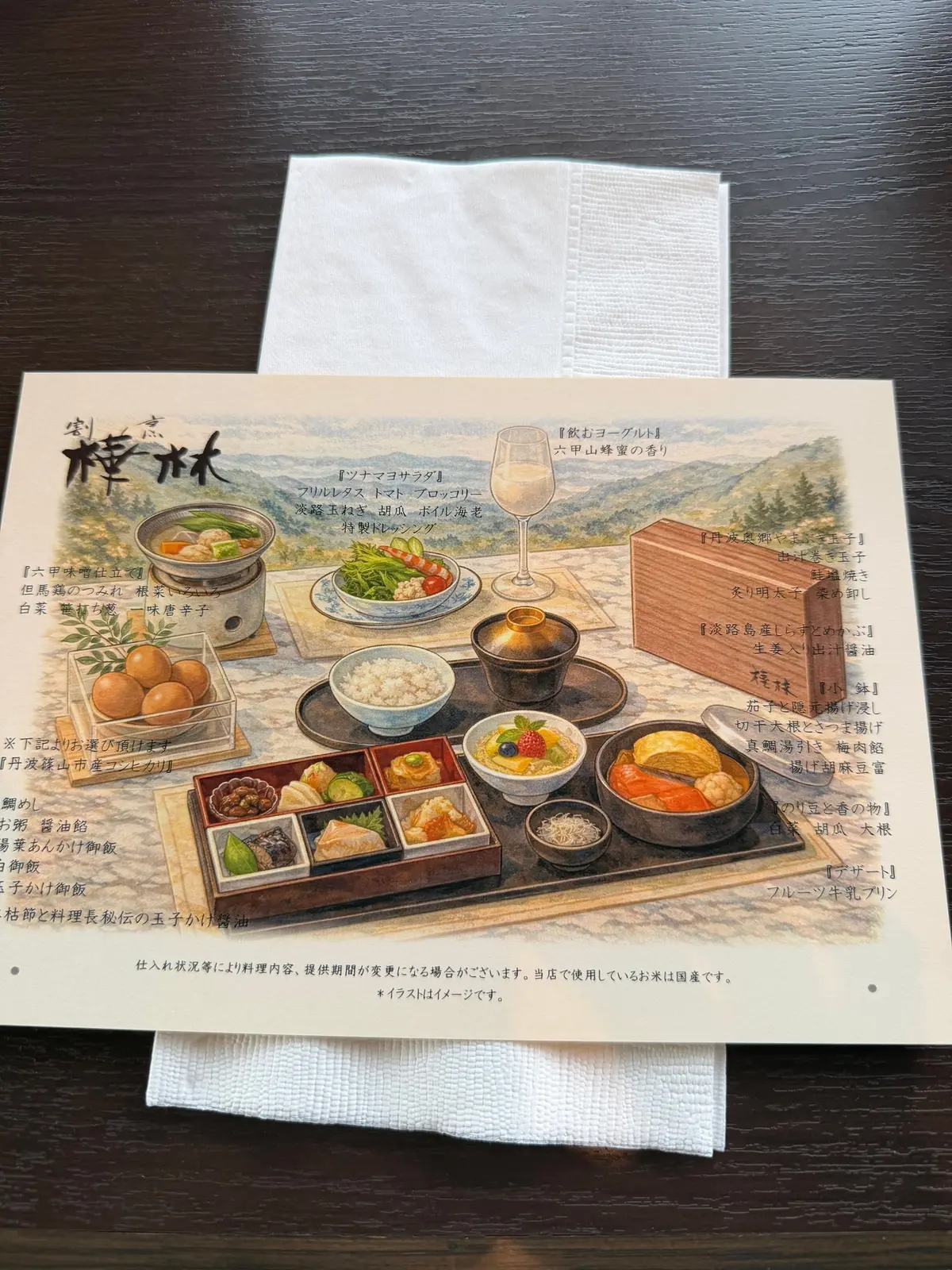木目調のテーブルに置かれた料理の写真。中央には、複数の小皿に盛り付けられた和食料理が並び、手前には湯気の立つお茶碗と汁椀が置かれている。右側には、お箸と茶碗が配置されている。背景には、料理名が書かれたメニューが貼られている。