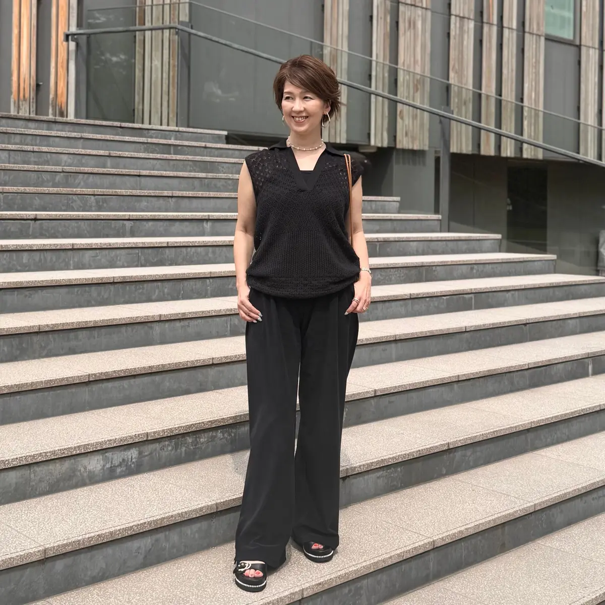 黒いニットのタンクトップと黒いワイドパンツを着用した女性が、灰色の階段に立って微笑んでいる。茶色のボブカットで、右手に手を当てている。背景には現代的な建築物が見える。