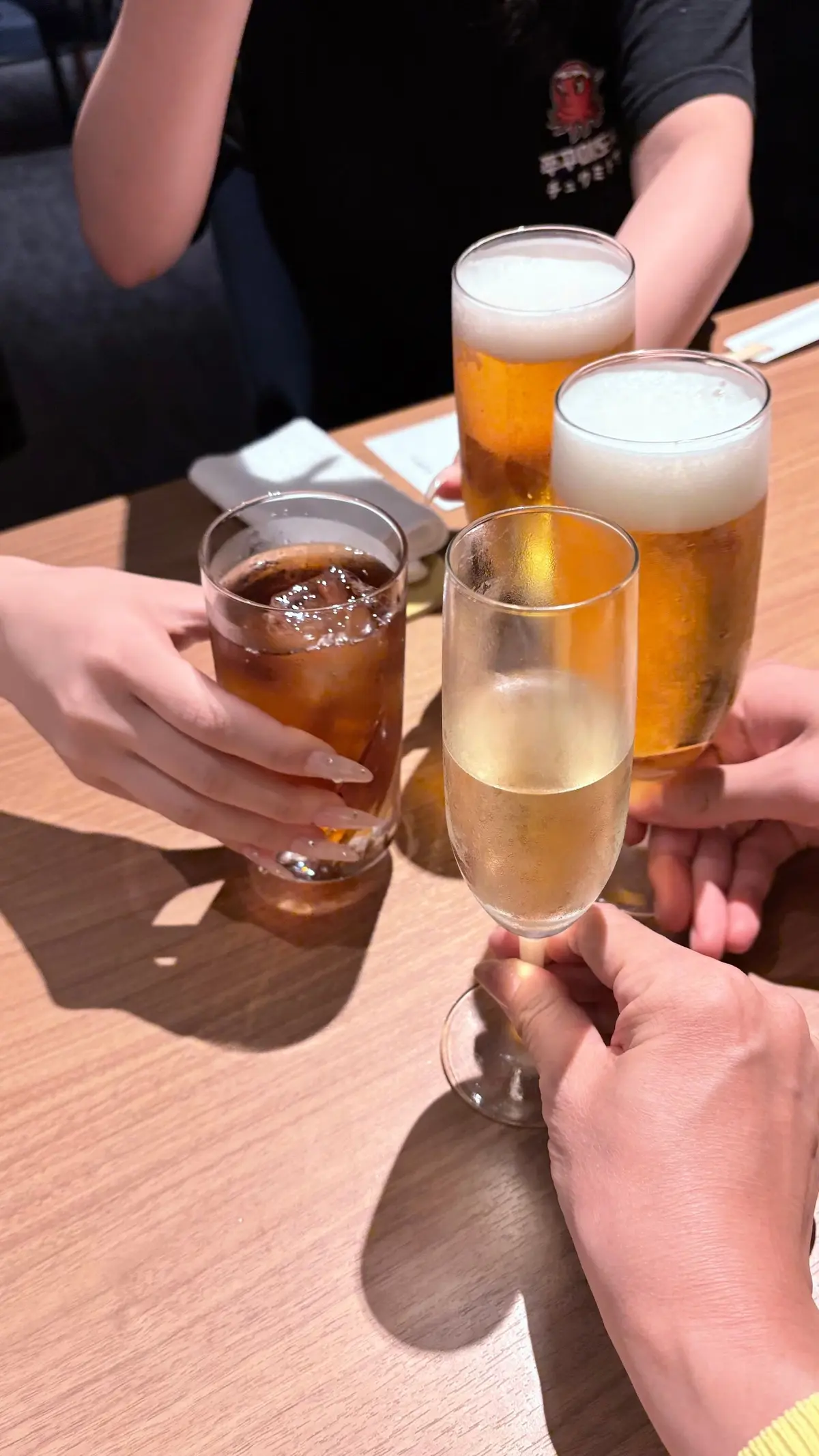 木製のテーブルに置かれた、様々な飲み物が並んでいる様子。左から、アイスコーヒー、ビール、シャンパンのグラスが、人々の手によって軽く握られている。背景には、黒いテーブルクロスと人物の姿が見える。