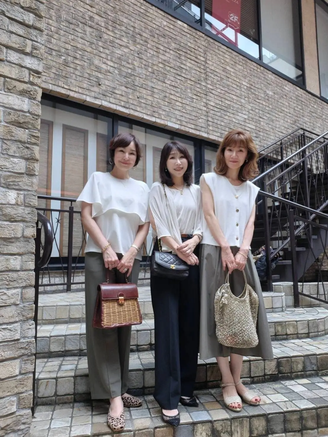 ベージュ系の服装をした3人の女性が、石造りの建物の前で並んで立っている。左から、白Tシャツにベージュのパンツ、ウィッカーのバッグを持つ女性。中央は、白のブラウスに黒のパンツ、黒いチェーンバッグを持つ女性。
