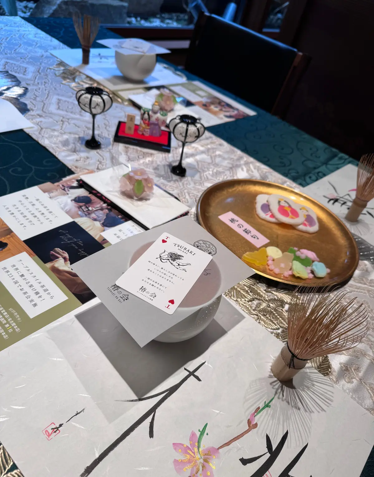 テーブルに並べられた、椿（ツバキ）の和風お菓子と飲み物。木製の托（と）に抹茶のお菓子、小皿に白玉、金色の小皿に赤い餡が入ったお菓子。テーブルクロスは緑色の葉柄、紙には日本語のテキストとハート形のマーク。
