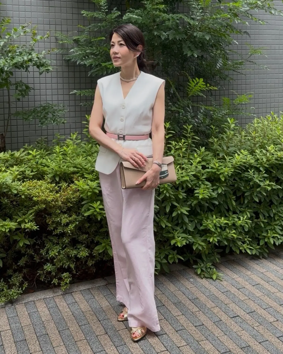 女性が白の無袖ジャケットとピンクのワイドパンツを着用し、ピンクのベルトでウエストを締めています。左手には茶色のクラッチバッグを持ち、背景には緑の低木が並び、グレーのタイル壁が映っています。