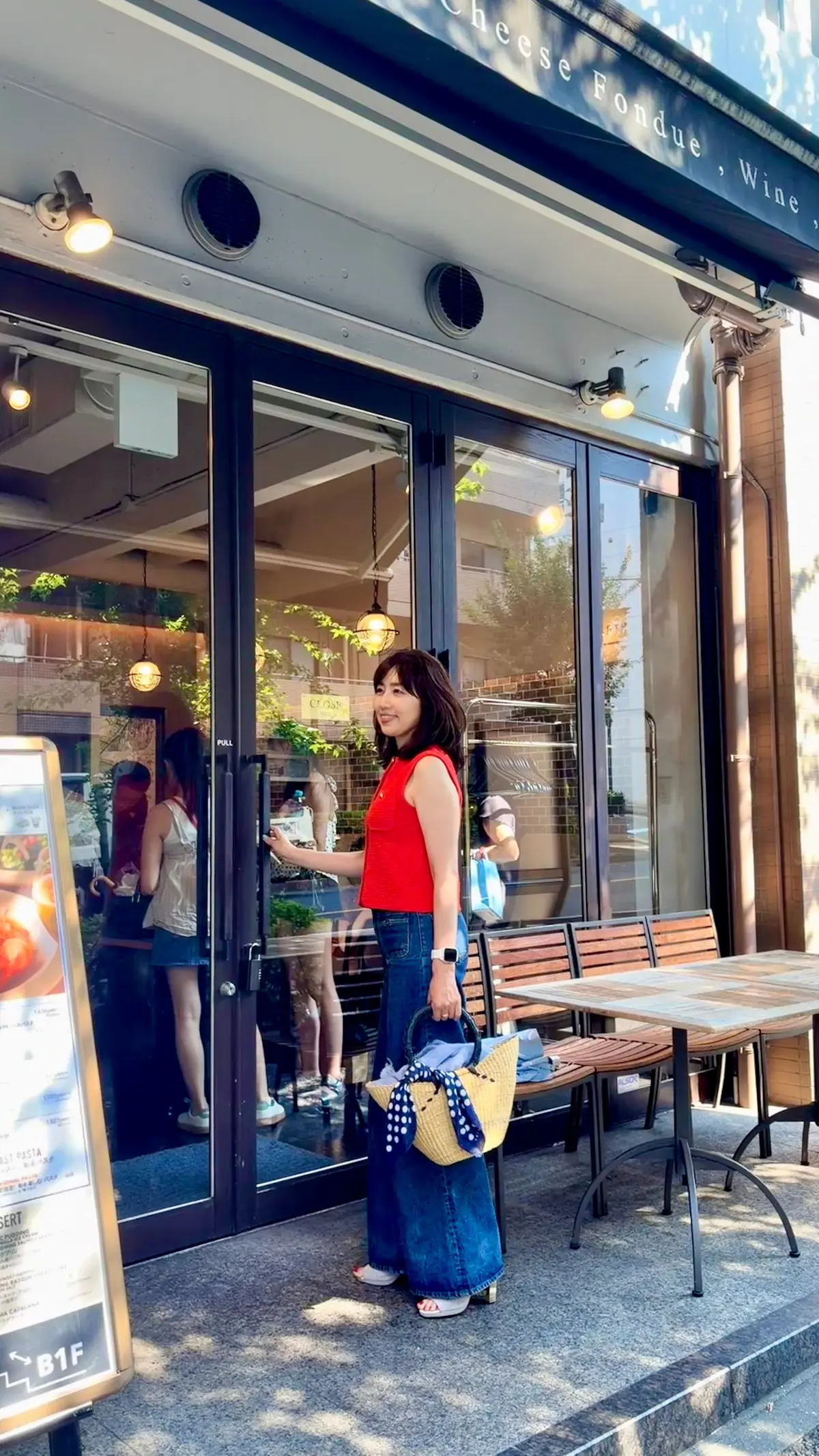 女性がレストランの入り口に立っている写真。茶色の髪をポニーテールにし、黒いスキニーデニムと白いトップスを着用。右手に麦わら帽子と青いバッグを持っている。背景には、チーズフォンデュとワインを提供するレストランの窓とテーブルセットが見える。