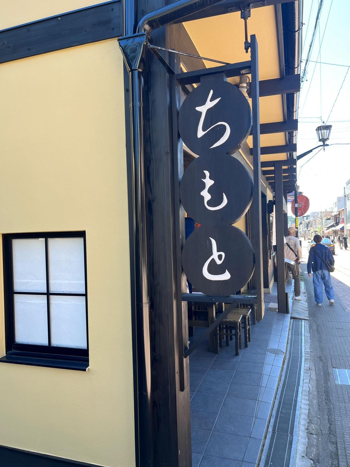 黄色い壁の建物に設置された、黒い円形の看板が縦に3枚並んでいる。看板には白い文字で「ちもと」と書かれている。看板の奥には、テーブルと椅子が並び、歩道には人々が歩いている。