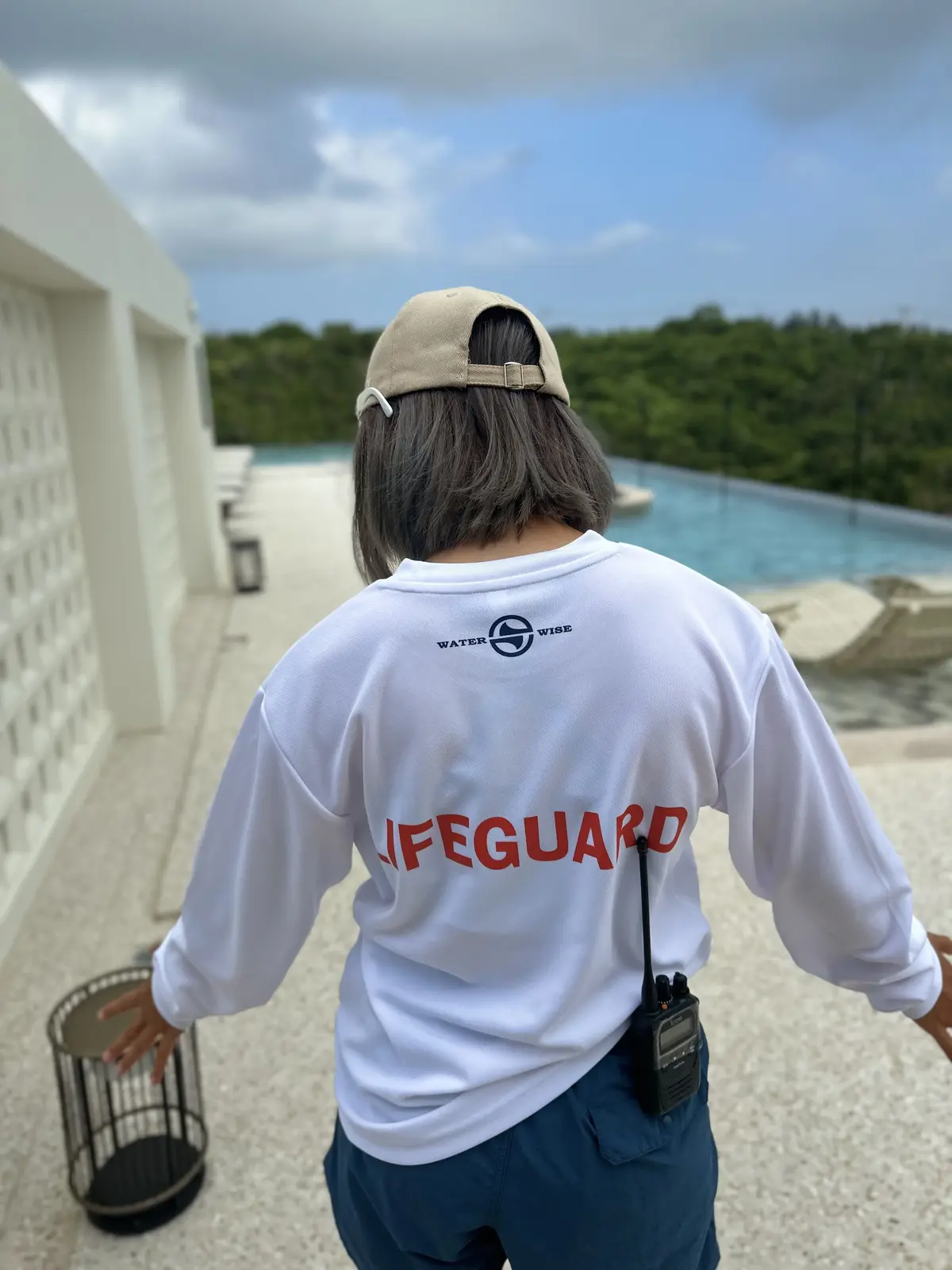 白い長袖シャツを着たライフガード。背中には「WATER WISE」のロゴと赤い「Lifeguard」の文字。キャップを被り、無線機を装着。背景にはプールと緑の木々。