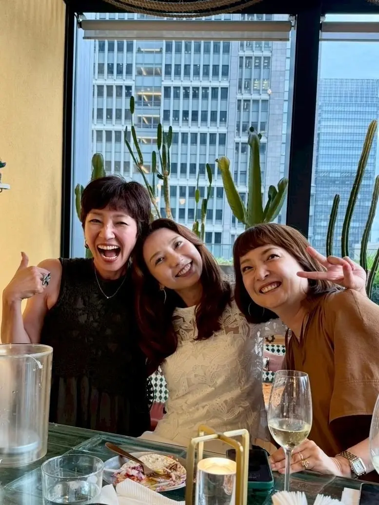 3人の女性が笑顔で写る屋内写真。窓の外には高層ビル群が広がり、室内には観葉植物が置かれている。テーブルにはワイングラスや料理が並び、楽しげな雰囲気が伝わる。
