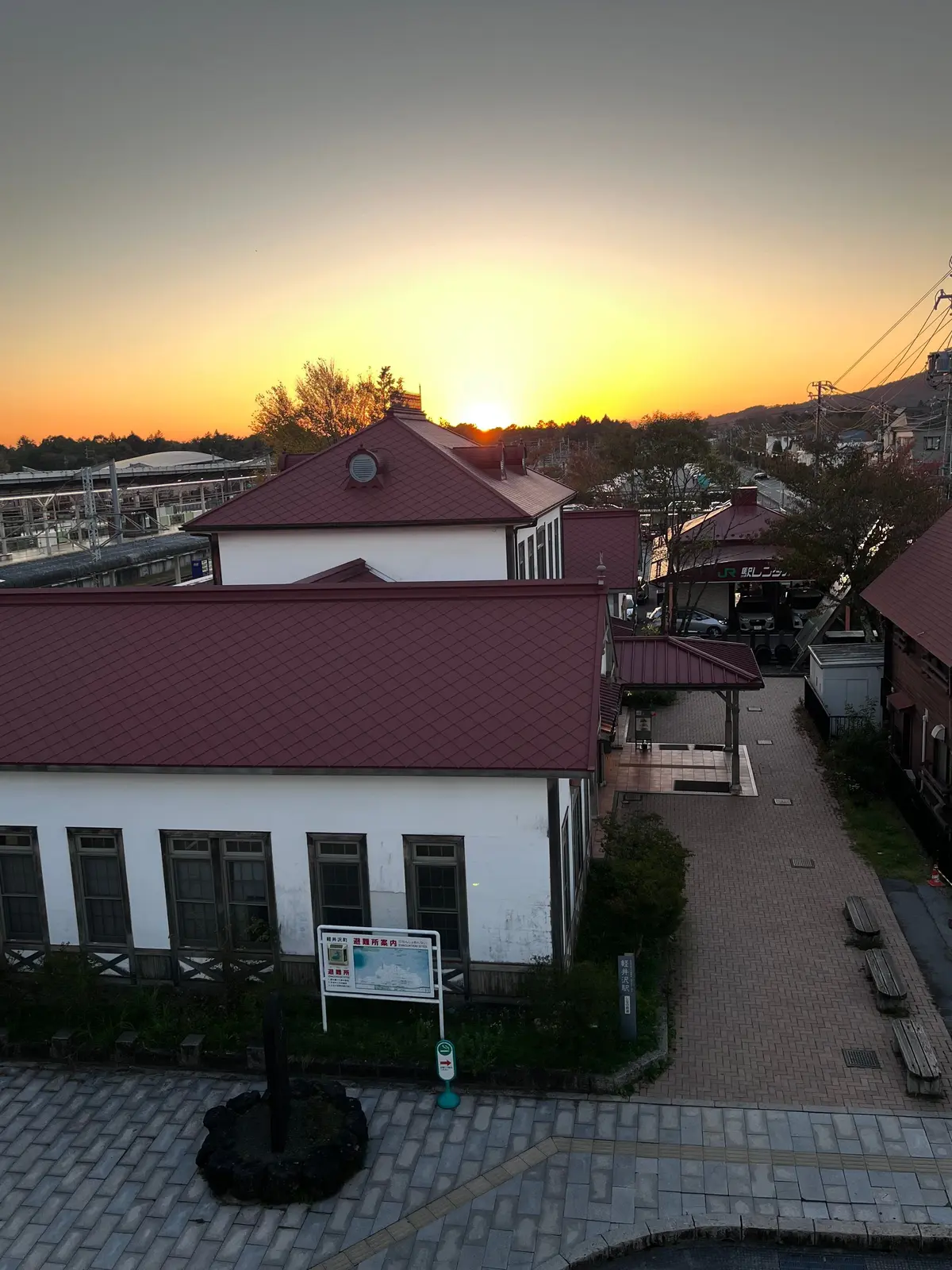 夕焼け空の下、赤い屋根の古い駅舎と周辺の風景。駅の前に案内板があり、歩道にはベンチが並ぶ。背景には、住宅街と山並みが広がる。