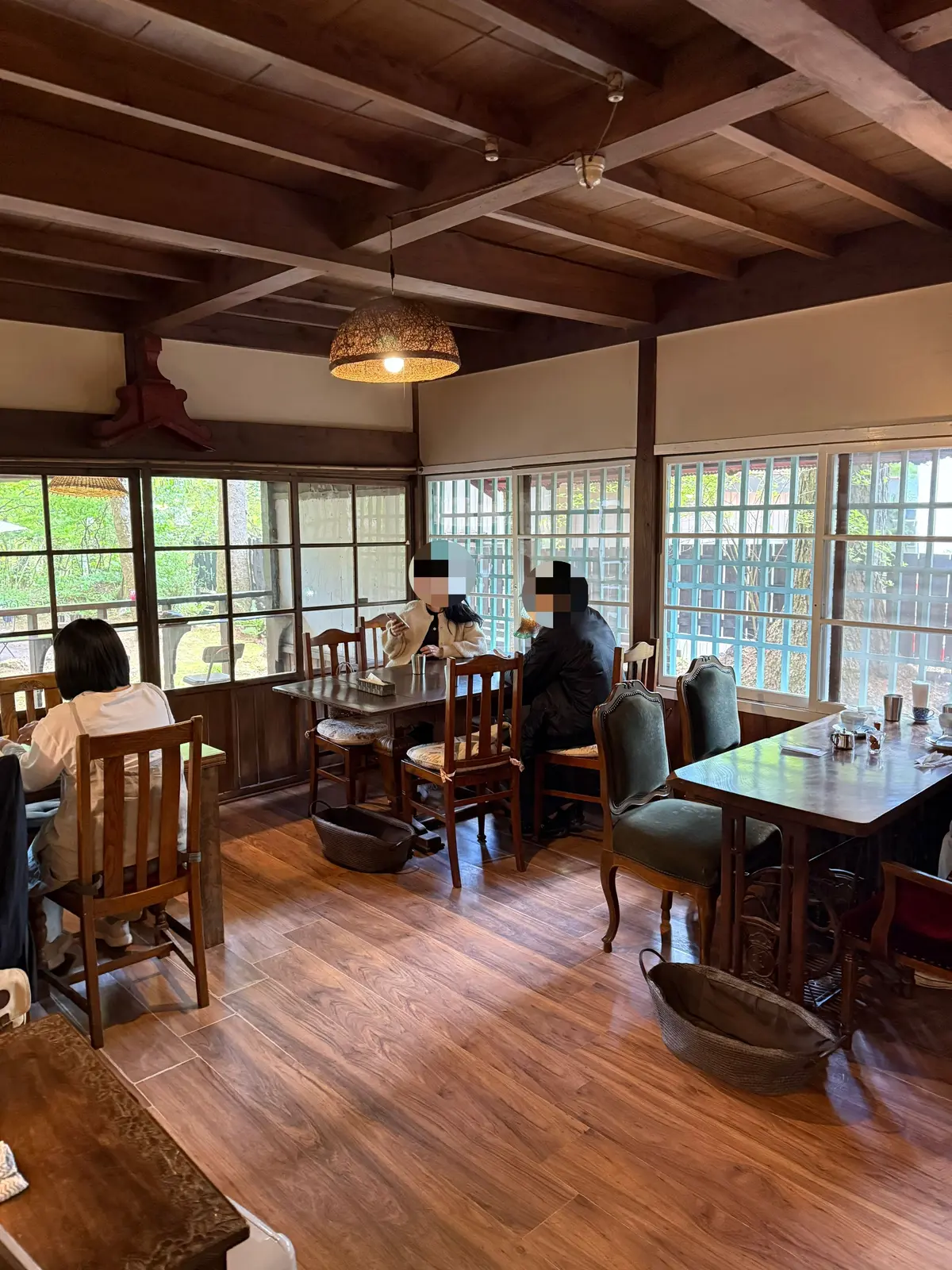 木製の天井と床の落ち着いたカフェで、窓から自然光が差し込む。テーブルと椅子が配置され、客人が食事を楽しんでいる。中央に編み込まれた照明が温かな光を放ち、籐のバスケットが床に置かれている。