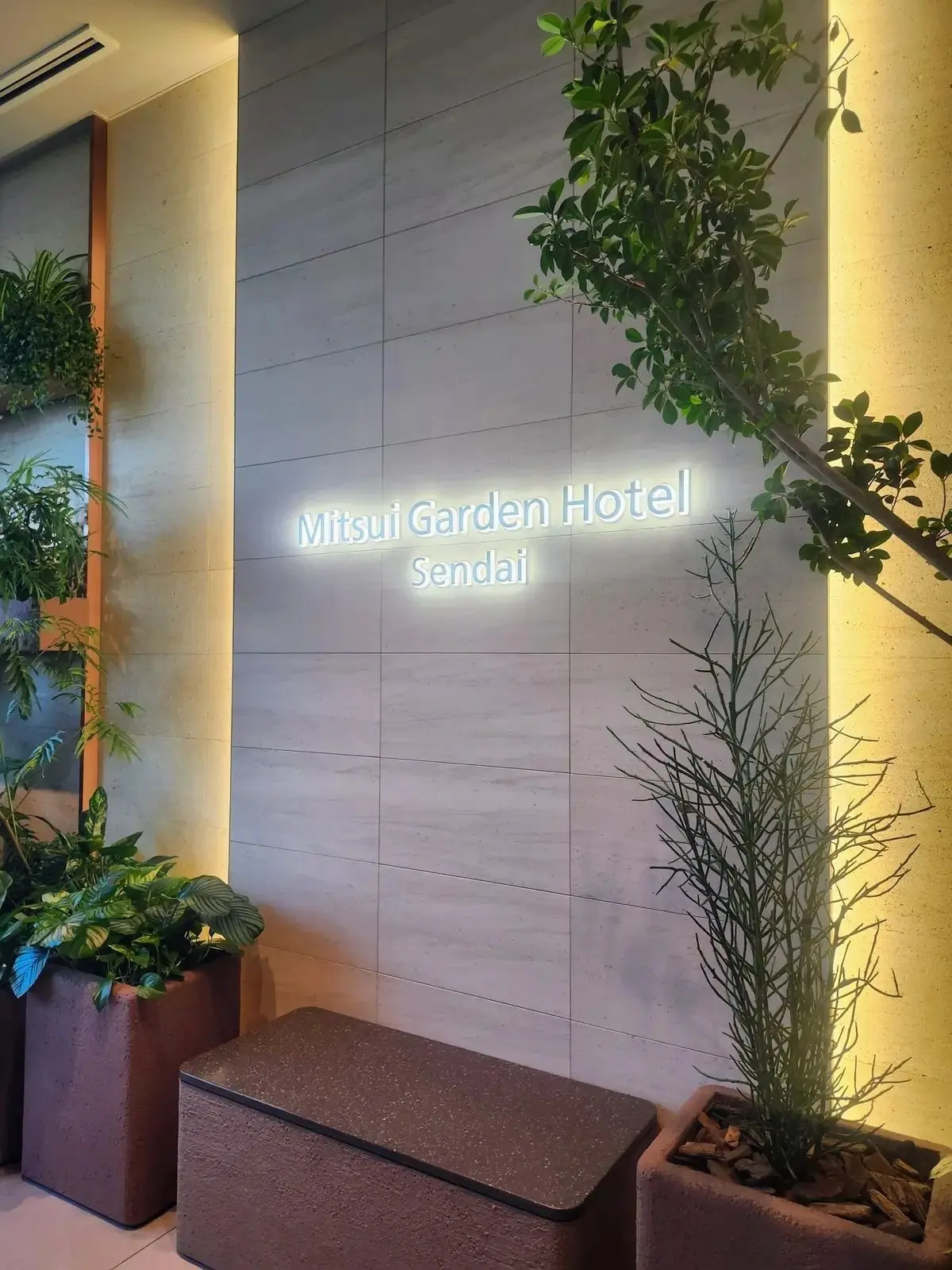 グレーのタイル壁に、光る「Mitsui Garden Hotel Sendai」のサイン。左右に植栽があり、右側には葉の多い木と細い草木が置かれている。足元には灰色の収納ボックス。