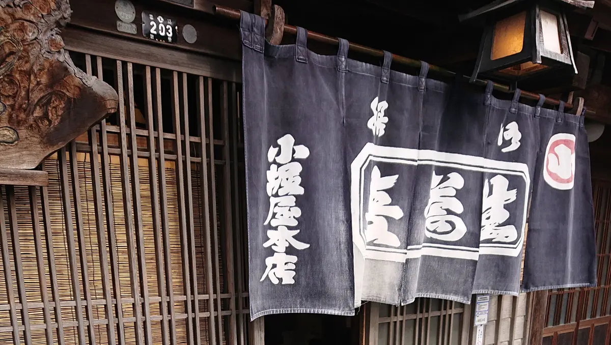 伝統的な木造建築の入り口前に立つ少年。黒い帽子とマスクを着用し、車が描かれた白いTシャツと茶色のジャケットを着ている。手にはリュックを背負っている。入り口には「小笹屋本店」の看板が掛かっている。