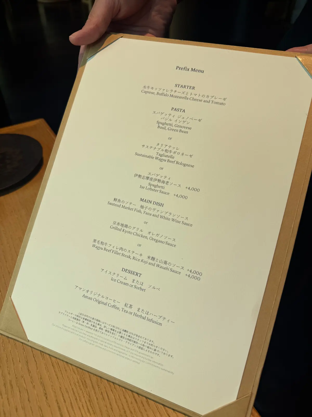 レストランのプレフィックスメニュー。左上に「Prefix Menu」の文字、下には日本語の料理名と価格が記載されている。背景は木目調のテーブルクロス。京都のラグジュアリーリゾートホテル、アマン京都。AMAN京都。ザ・リビング パビリオン by アマン。ランチタイム、京都観光。京都ホテル。