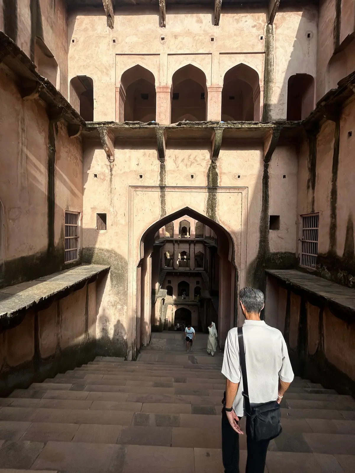 インドの古城の石段を上る男性。左右にアーチ型の窓が並び、奥にはさらに階段と建物が続く。壁はピンク色で、光と影がコントラストを生み出している。