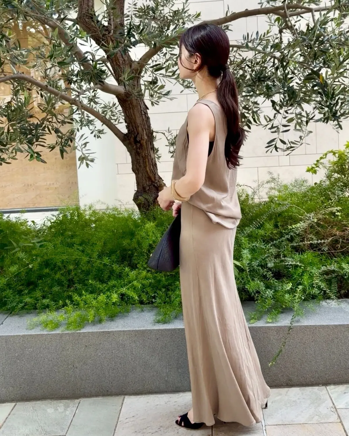 ベージュのトップスとロングスカートのセットアップを着た女性が、屋外の石畳の道に立っている。後ろ姿で、右手に黒いバッグを持ち、左手でスカートの裾を持つ。背景には緑の植栽と白い壁、そして木が見える。