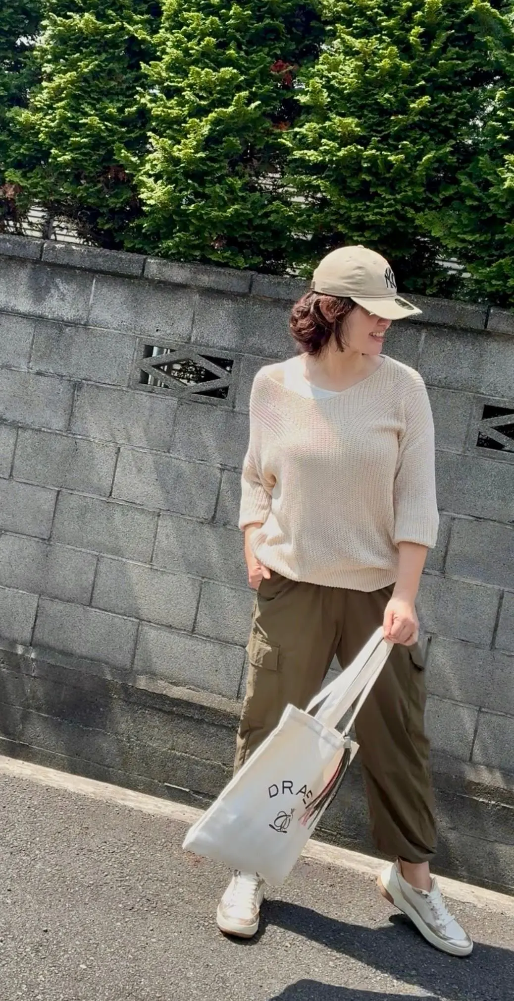 ベージュのキャップを被った女性が、白いニットとカーゴパンツ、白いスニーカーを着用し、白いプラダのトートバッグを持って歩いている。背景にはコンクリートの壁と緑の木々が見える。
