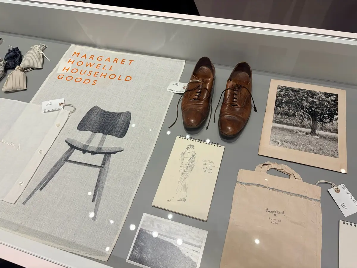 展示ケース内の商品群。左上には「MARGARET HOWELL HOUSEHOLD GOODS」と表記された書籍、中央には木製椅子と靴、右には白黒写真、下部にはバッグやノートなどが並んでいる。全体的に落ち着いた色合いで、上品な雰囲気が伝わる。