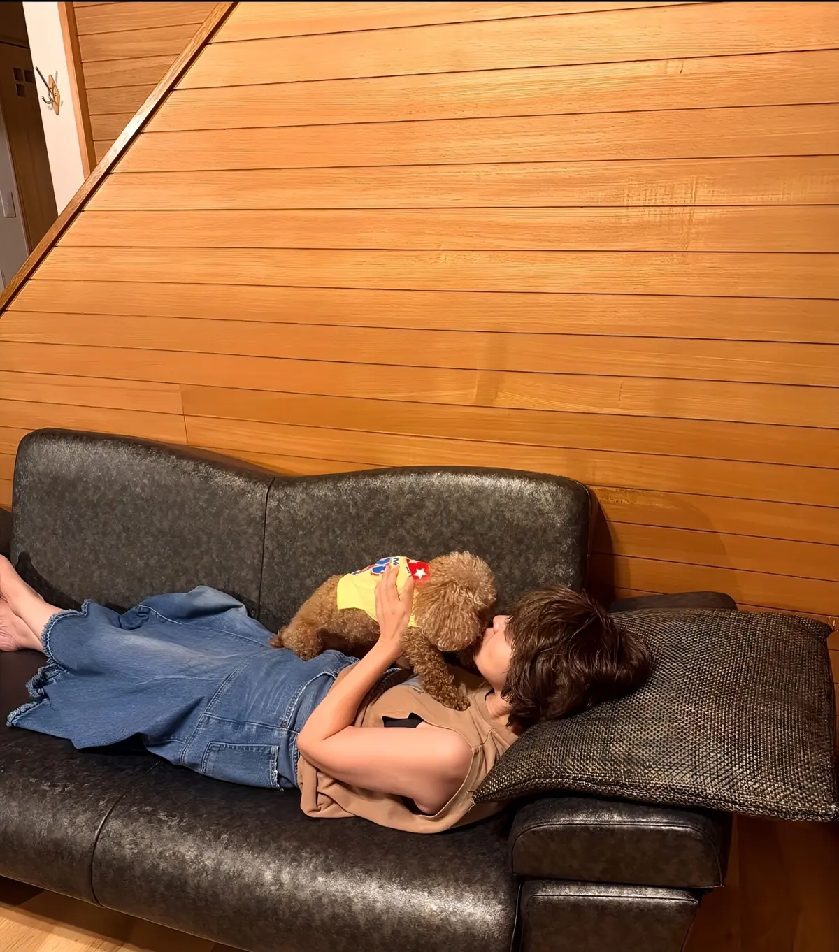 ソファでくつろぐ若い女性と、寄り添うように眠る白いプードルの写真。女性はデニムのオーバーオールと黒いTシャツを着ており、片足はソファからぶら下がっている。背景には木目調の壁がある。