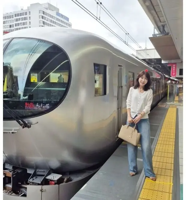駅のホームに立つ女性が、銀色の新幹線車両の横で微笑んでいる。女性は白いシャツにジーンズ、ベージュのハンドバッグを身につけている。車両の前面は丸みを帯びており、大きな窓がある。背景には高層ビルと駅の屋根が見える。