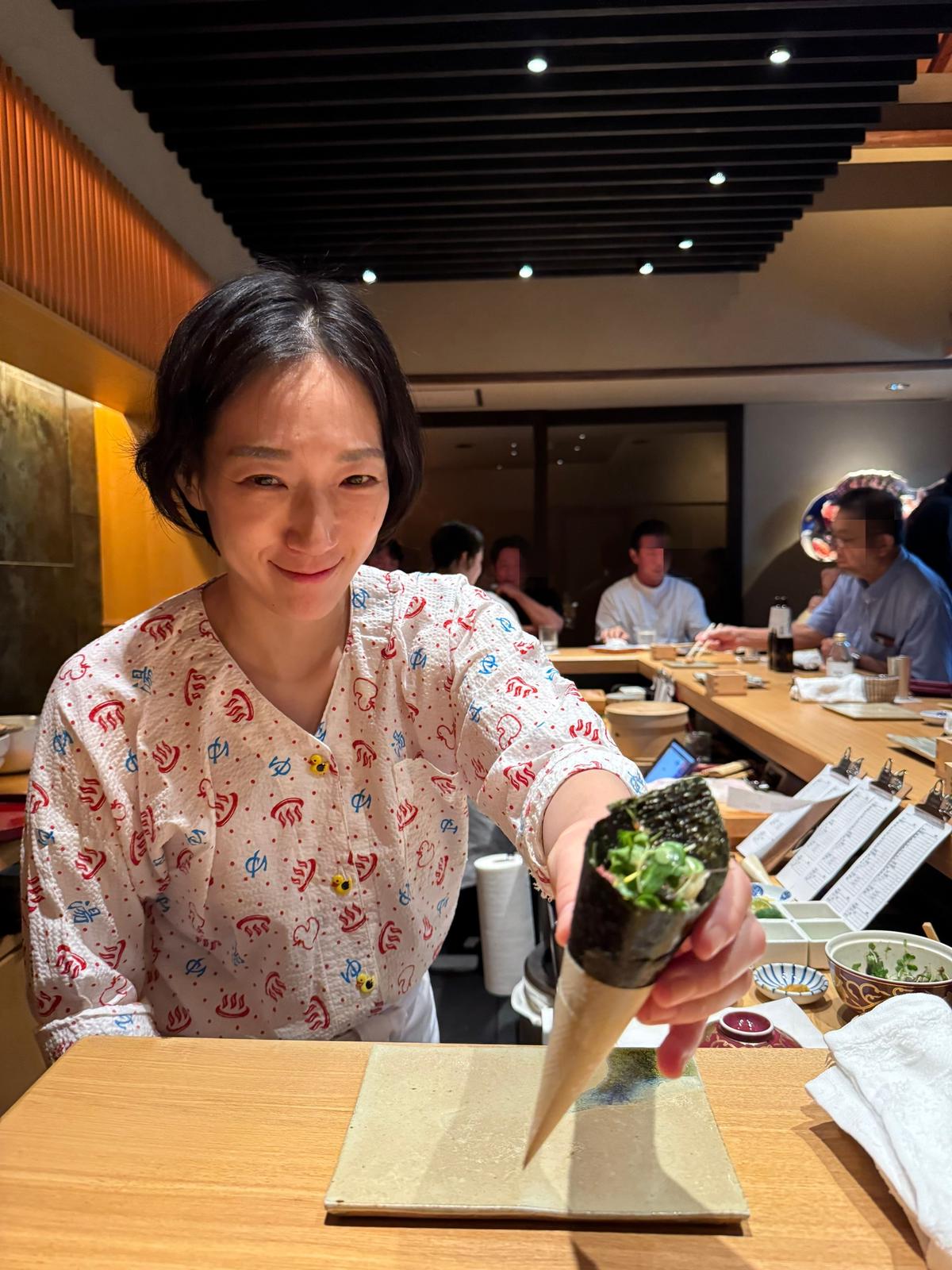 笑顔の女性が、手で握った海苔巻きに粉をふりかけている。背景には、他の客が食事をする寿司屋の店内が見える。