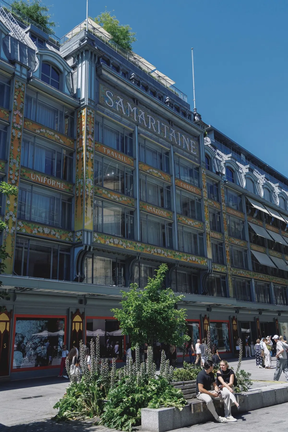 パリの老舗百貨店サマリテーヌの外観。黄色の装飾が施された建物で、窓には商品名が掲示されている。正面には植栽スペースがあり、人々が休憩している。