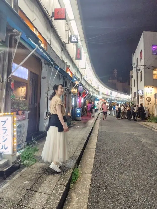 夜の商店街で、女性が右手を前に出してこちらを見ている。白いチュニックと黒いスカート、白いスニーカーを着用。商店街には提灯や看板が並び、多くの人が歩いている。