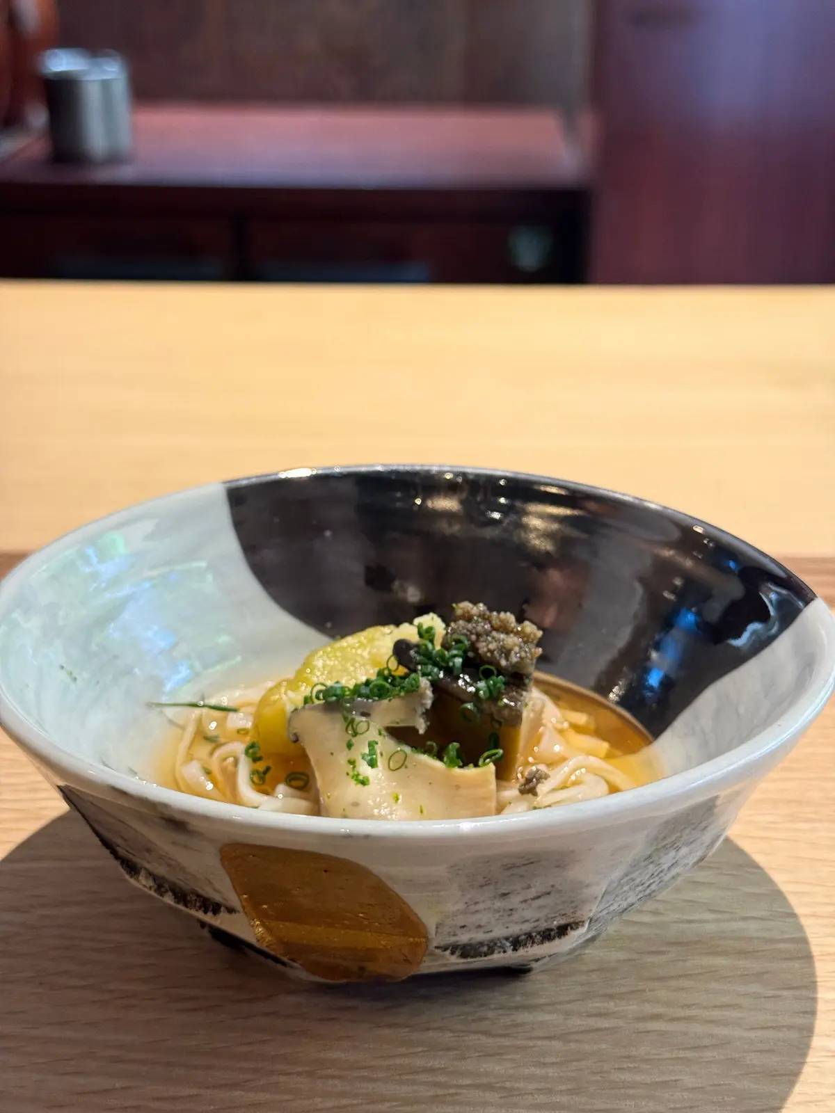 木製のテーブルに置かれた、模様入りの陶器の器に入った料理。うどんのような麺と、豆腐、柑橘系のスライス、ハーブ、キノコなどが、茶色のスープで提供されている。