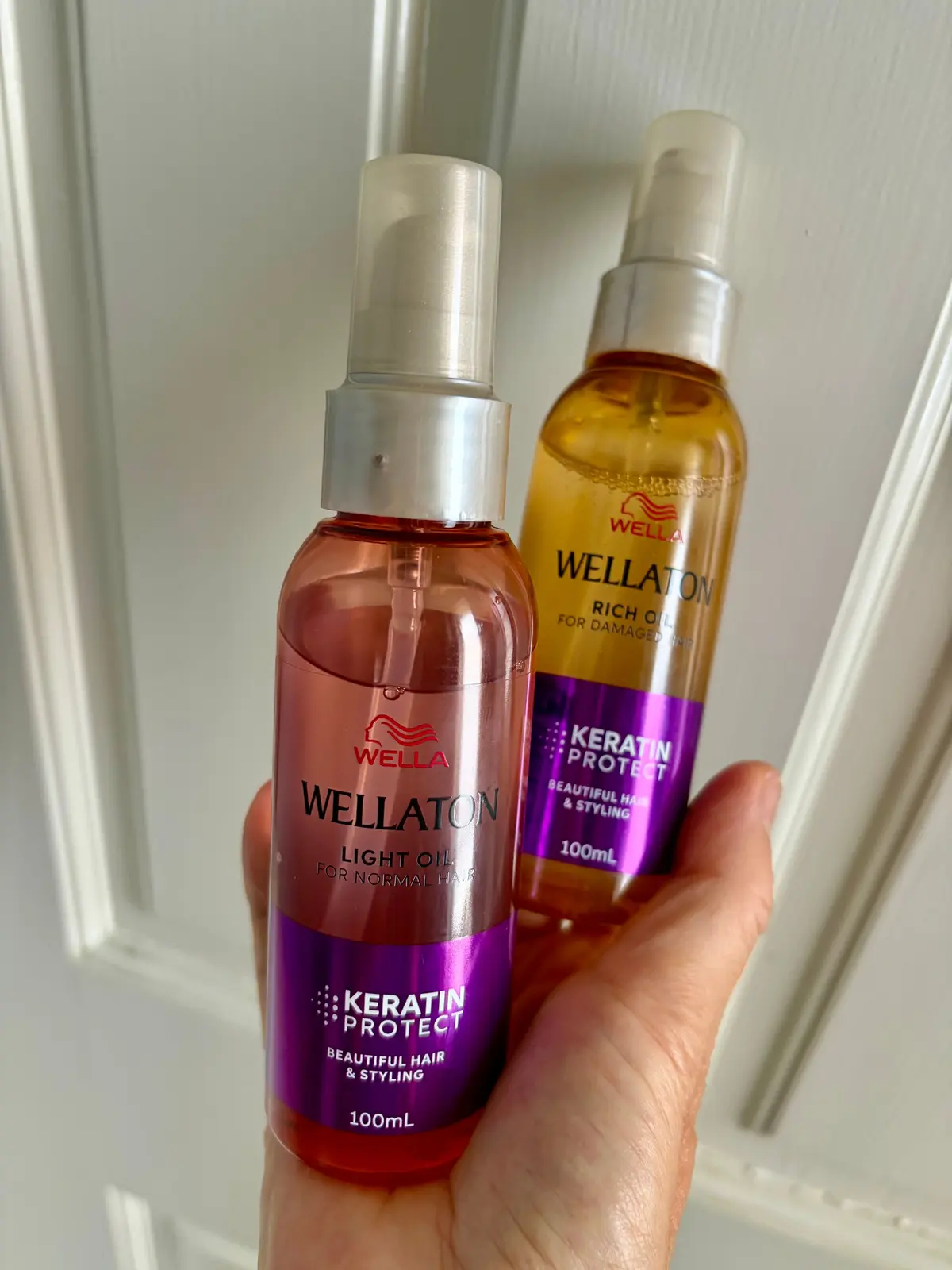 手に持たれた2本のウェラ（Wella）のヘアオイル。左はピンク色のボトルで「ライトオイル」、右は金色のボトルで「リッチオイル」。どちらも100mlで、ケラチンプロテクトの文字が記載されている。背景は白い壁。