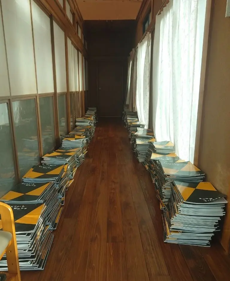 長方形の書籍が両側に積み上げられた、木製の廊下。窓から光が差し込み、書籍の黄色と黒の模様が映える。奥は暗く、奥行きを感じさせる。