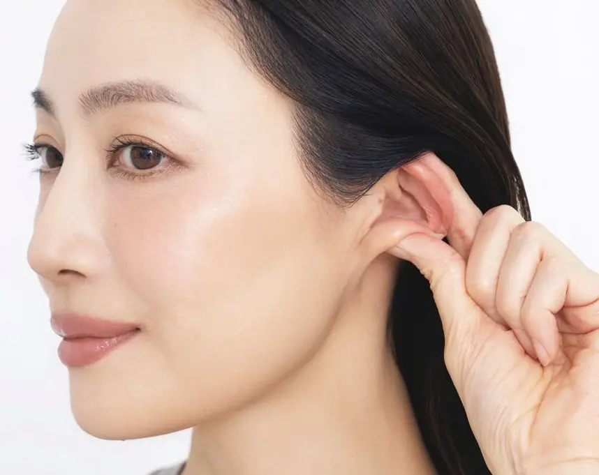 耳をつまんで、耳まわりに 多くあるツボを刺激　女性が耳をふくらませ、耳垢を取り除こうとしている。白い背景。