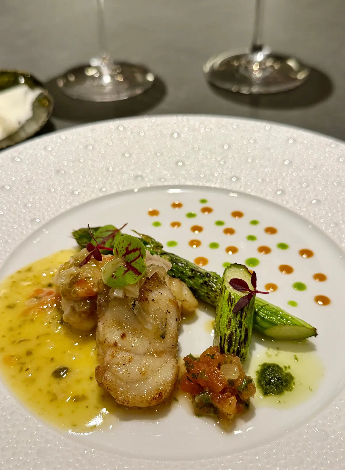 白磁の皿に盛り付けられた、魚料理の盛り合わせ。魚のソテー、グリルしたアスパラガス、トマトの具材、ソースが配置されている。赤紫色の葉が添えられ、全体的に上品な印象。