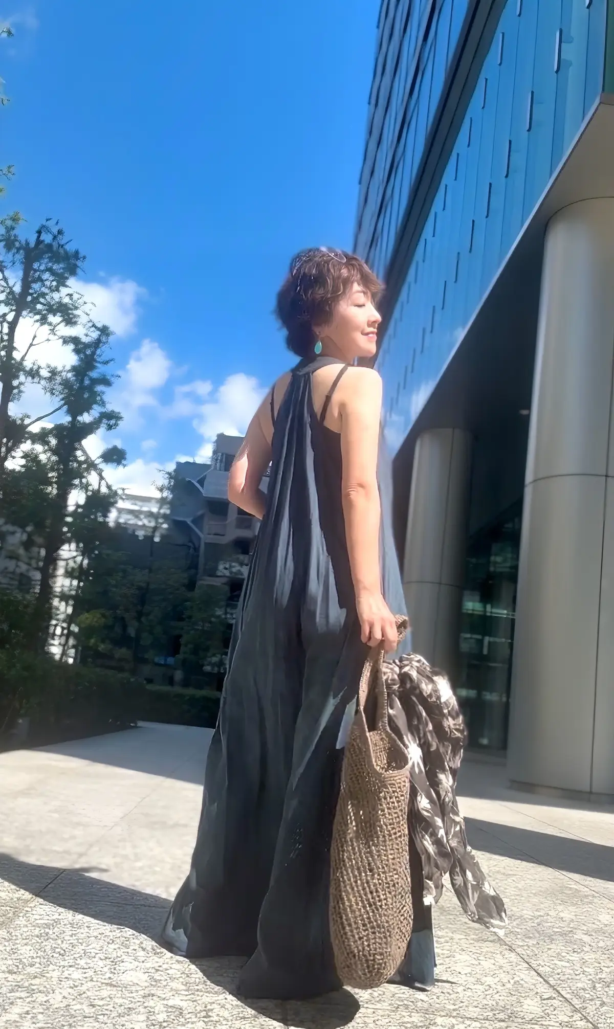 黒いスリップドレスを着た女性が、建物のそばで振り返り微笑んでいる。右手に編み物のバッグを持ち、背景には青空と緑の木々が見える。