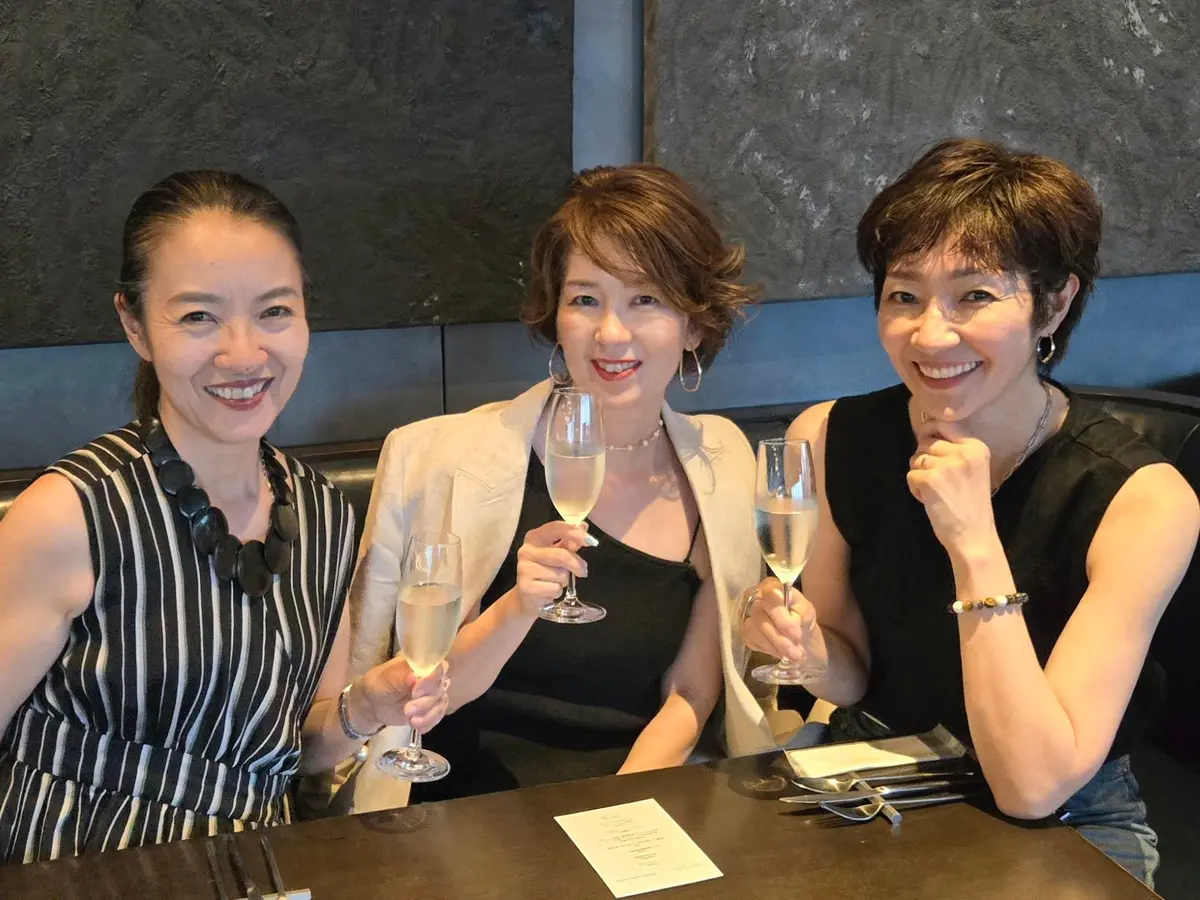 黒いテーブル席に座り、シャンパングラスを持つ3人のアジア系の女性。左と右の女性は黒い服、中央の女性は黒いドレスに白いジャケットを着用。笑顔でこちらを見ている。背景はグレーの壁。