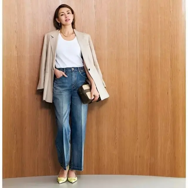 女性が木製の壁に寄りかかり、ベージュのコートとデニムパンツを着て、小さな手袋を持っている